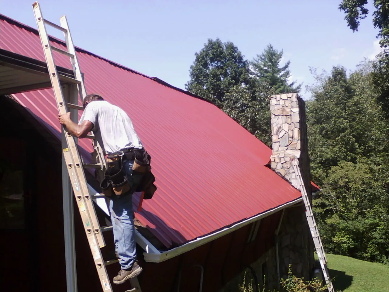 Roofer on a ladder beside a red metal roof and stone chimney — Soffit & Fascia Repair by Roofing Brothers in Kiowa